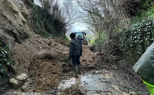 A landslide in Barrington, Ilminster with a person standing in wellies in the foreground and a tractor in the background approaching the landslide[