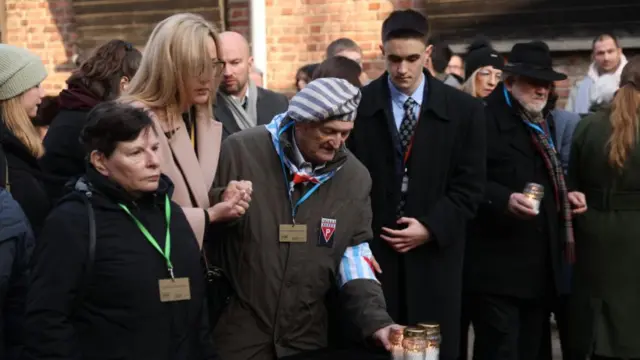 A crowd of people gathered at Auschwitz including one man wearing a blue and white striped hat, who reaches for a candle