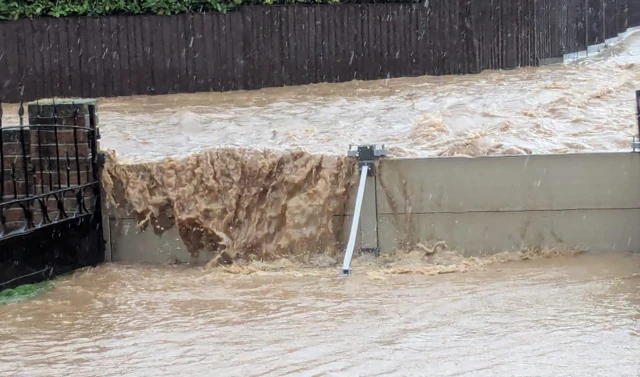 Flood water goes over a barrier.  The water is muddy and is running down the street behind the barrier