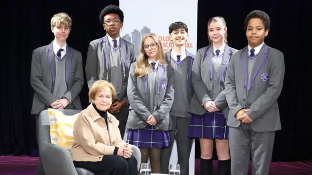 Mala Tribich, seated, with six young pupils standing near her in school uniform, all looking at the camera