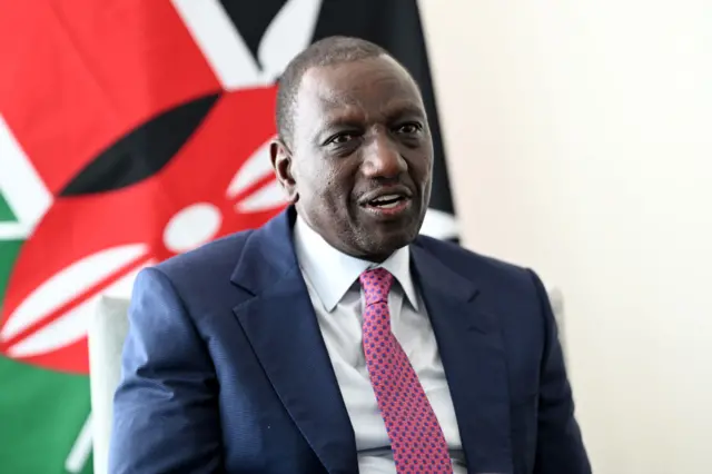 William Ruto, wearing a blue suit and multi-coloured tie, sits in front of a Kenyan flag