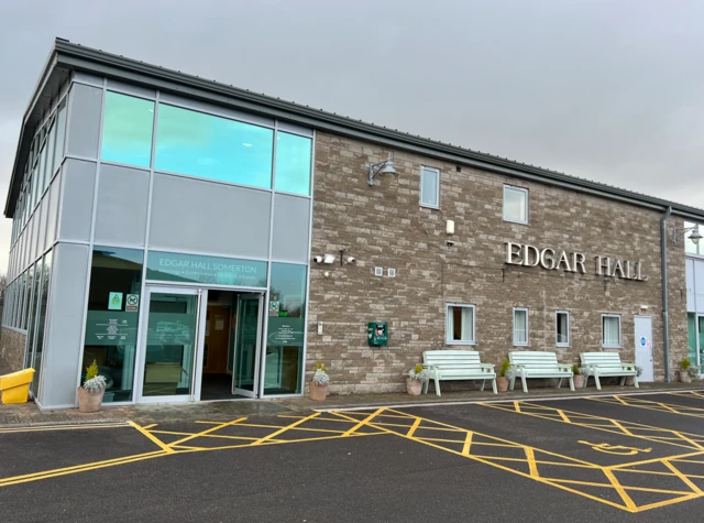 The picture shows the outside of Edgar Hall in Somerton. There is brick wall and double glass doors. Benches and plant pots are on the walls outside