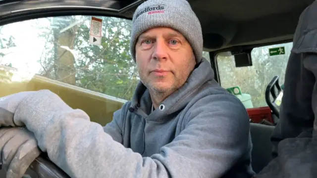 Graham wearing a grey hat and hoodie and grey gloves at the wheel of his telehandler