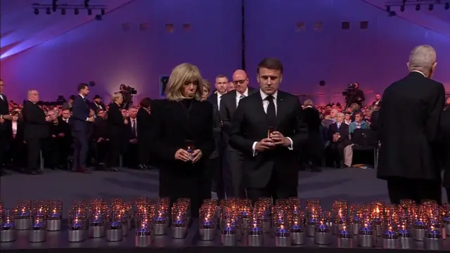 Emmanuel Macron and Brigitte Macron each hold a candle wearing black. The stand in front of dozens of candles