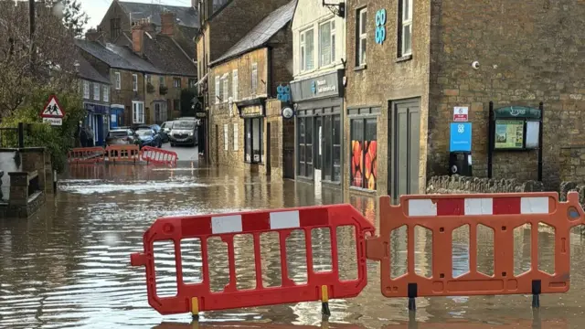 Red barriers prevent access to a flooded road with some shop fronts which are inaccessible.