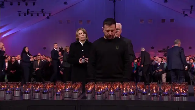 Volodymyr Zelensky bows his head in front of dozens of candles. Rows of people are seated behind him.