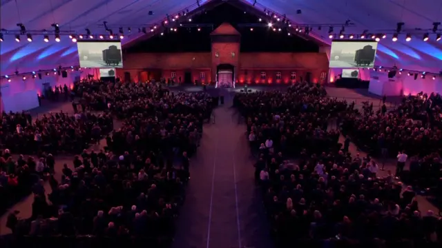 Large white tent with rows of seating and people sat down in large audience looking at stage