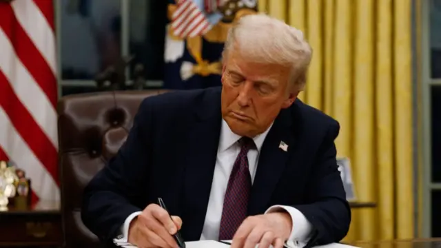 Donald Trump sits at a desk while he looks down to sign an executive order