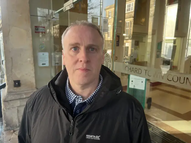 Councillor Jason Baker is looking into the camera. He is wearing a dark jacket and is standing in front of the Guildhall