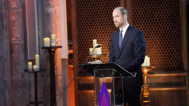 William, wearing a suit, stands at a lectern decorated with a purple flame