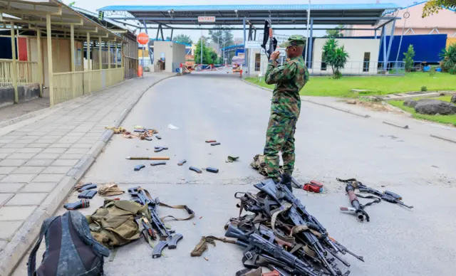 A soldier wearing military uniform inspects weapons scattered on the floor.