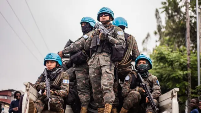 UN soldiers wearing military fatigues and blue helmets ride at the back of a pickup truck. They carry guns.