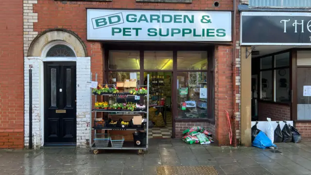 The outside of BD Garden & Pet Supplies after flood water has drained away. The shop is on the bottom floor of a red brick building and has shelves of bedding flowers outside.