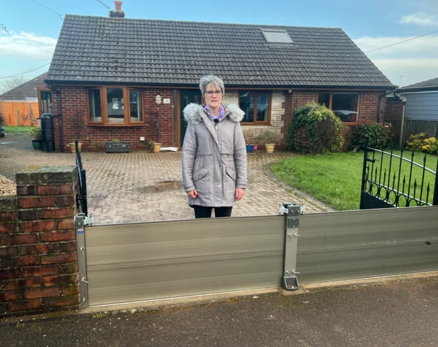 Elaine Merrick-Reed, in a grey coat with a fur-lined hood, stands behind a solid fence that sits in front of black metal gates across her drive way