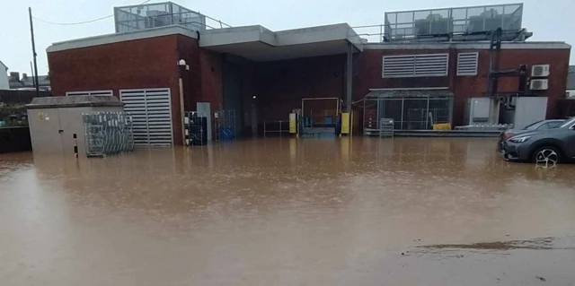 The picture shows the back of Sainsbury's in Chard with floodwater up to the trade entrance. Floodwater also surrounds parked cars next to the store.