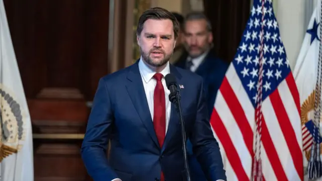 JD Vance wears a suit and speaks into a microphone. The American flag hangs behind him.