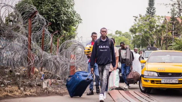 People walking with luggage like taxis next to an area guarded by barbed wire.