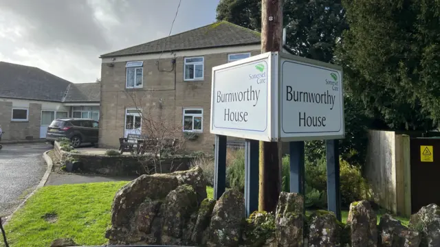 A sign which reads Burnworthy House on a white background, in front of the care home