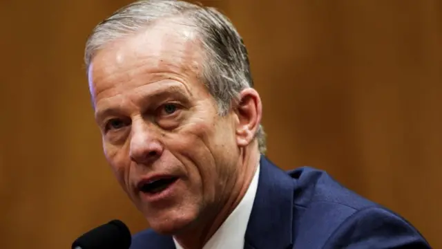 Republican Senate majority leader John Thune speaking into a microphone, seated. shot from shoulders up, wearing dark blue suit