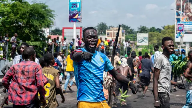 A protester in a blue top and yellow bottoms points past the camera as others gather during a demonstration