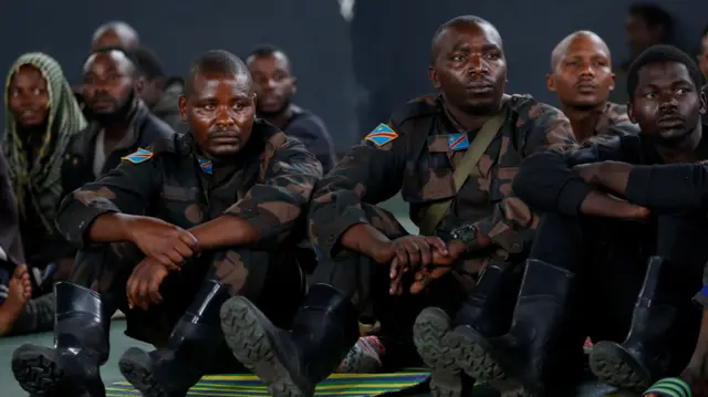 Men in military fatigues sit on the floor, looking expressionless