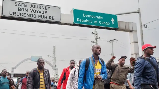 Refugees carrying belonging cross the border, passing underneath signs reading 'Democratic Republic of Congo' and 'Safe journey'