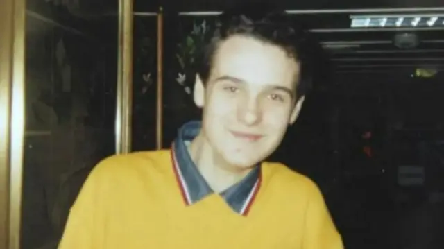 Alan Radford who has short brown curly hair, is wearing a yellow jumper with a blue shirt underneath that has a red and white collar