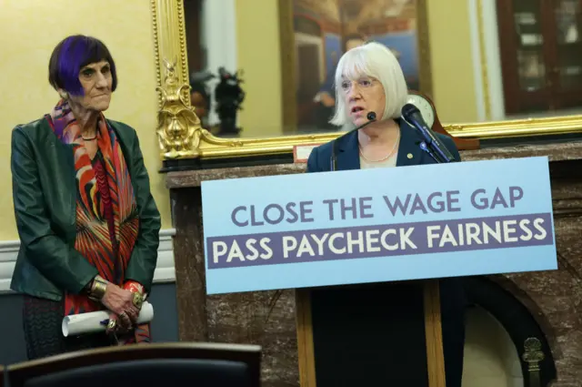 Two women stand at podium with a sign that reads "close the wage gap pass paycheck fairness"