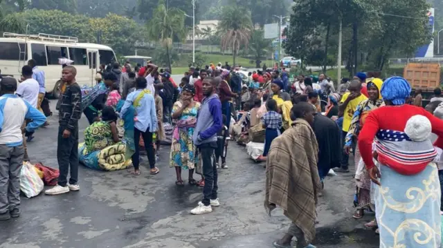 A group, carrying bedding and other belongings, gather outdoors, next to a white van.