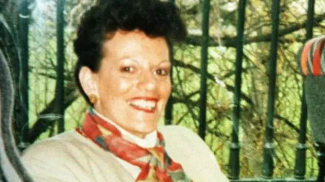 Ann McCombe has short brown hair, permed. She is wearing a cream jacket and a red,yellow and green scarf. She is sitting on a bus smiling at the camera.