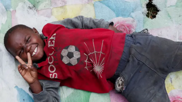 A Congolese child, having fled Goma for Rwanda, lies on a torn blanket smiles and throws up the 'peace' sign