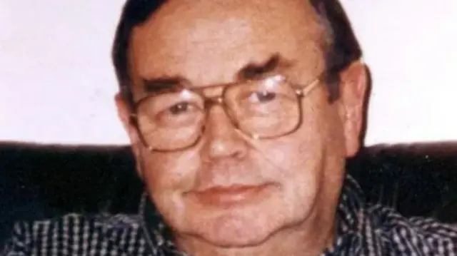 Sean McGrath pictured with short brown hair and wearing large gold framed glasses. He is wearing a checked black and white shirt.