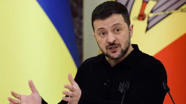 Ukraine's president Volodymyr Zelensky gesturing with both hands while speaking behind two microphones. Wearing a black long-sleeved top with partial Moldovan and Ukrainian flags in background