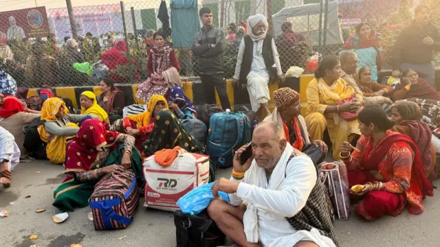 Crowds of people are seen on a roadway in front of a train station in Uttar Pradesh. They are wearing brightly coloured clothing, including saris, and are holding luggage.