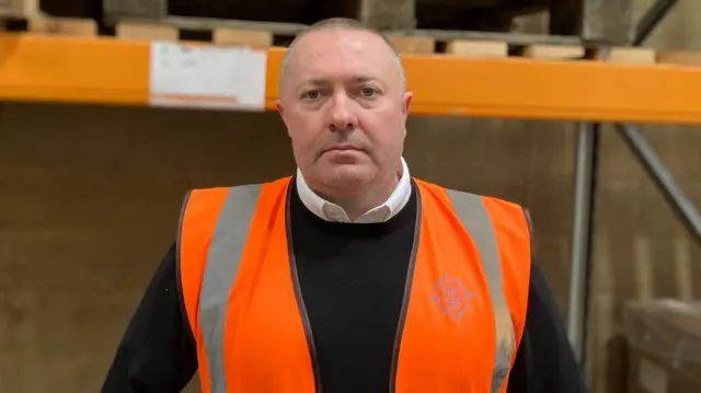 Mark Billany in a high vis vest over a white shirt and black jumper with scaffolding in the background