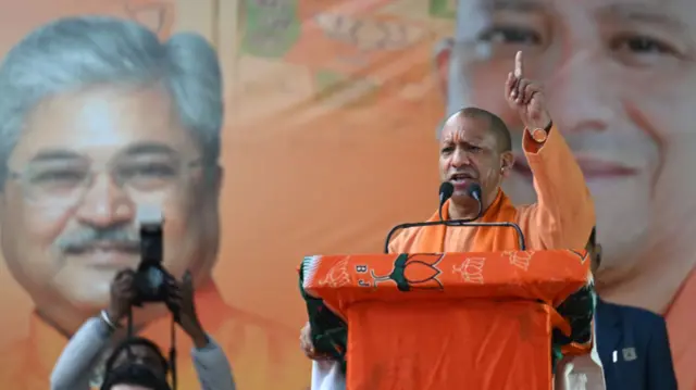 Yogi Adityanath wears orange, behind an orange podium, speaks with a microphone and points one finger in the air.