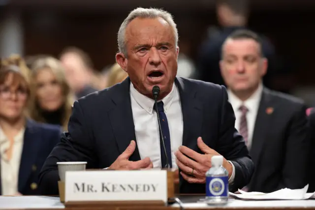 Robert F Kennedy Jr speaks at his hearing. His forehead his furrowed and his hands are raised to his chest. There's a placard with his name on it in front of him.