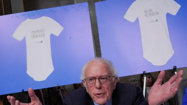 Bernie Sanders sits with arms raised and two posters behind him show children onesies with slogans "no vax no problem" and "Unvaxxed Unafraid"