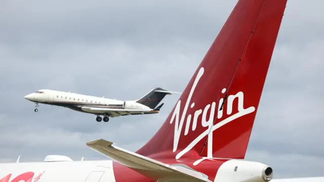 Tail fin of Virgin Atlantic aeroplane, with red background and white text, with a plane flying in the background