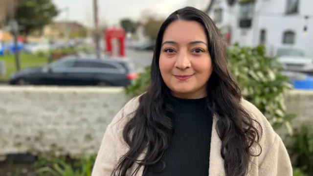 A woman with long brown hair and a brown jacket smiles for a picture.