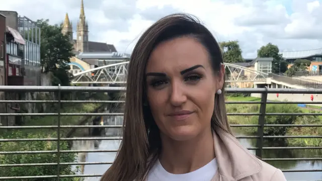 Claire Hayes with brown hair wearing a pink jacket and white top. She is standing on a bridge with a river and buildings behind