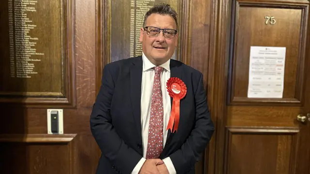 Karl Turner MP wearing a dark suit, floral tie and red Labour rosette stands in front of a dark wooden wall in Hull's Guildhall