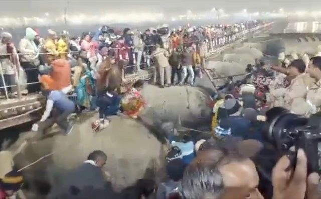 Crowds of people gather behind a fence with some falling as they attempt to reach the other side over the fence. Officers can be seen on the right.