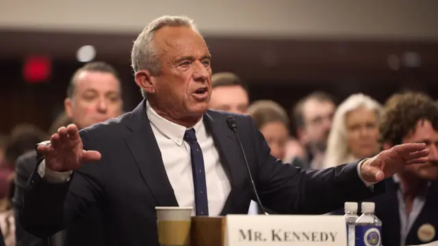 RFK Jr, seated, addresses the Senate committee while people watch on from behind