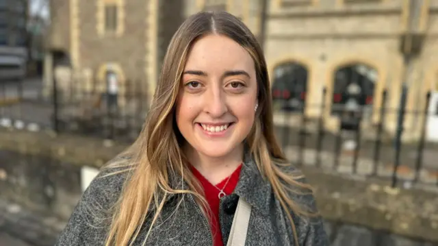 Isla Sutton smiling into camera, wearing grey coat and red jumper