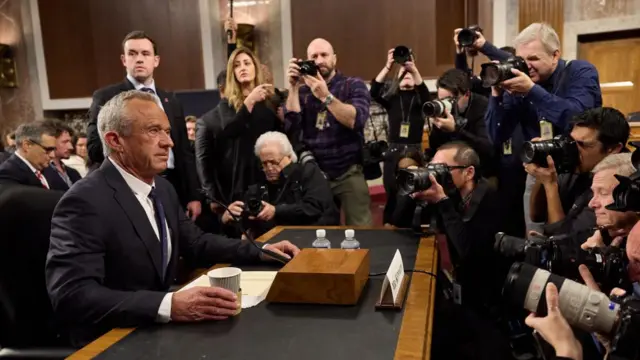 A side-view of Robert F Kennedy Jr sitting at his Senate hearing while surrounded by several photographers holding up their cameras.