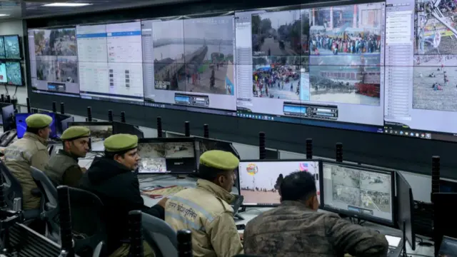 A row of police officers monitor two tiers of large screens showing CCTV footage of the Kumbh Mela