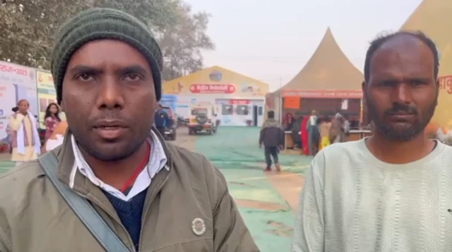 Two men standing side by side. One is wearing a green jacket and green hat, the other is wearing a white top