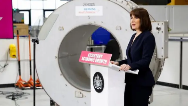 Rachel Reeves in navy blazer stood in a warehouse speaking behind a podium