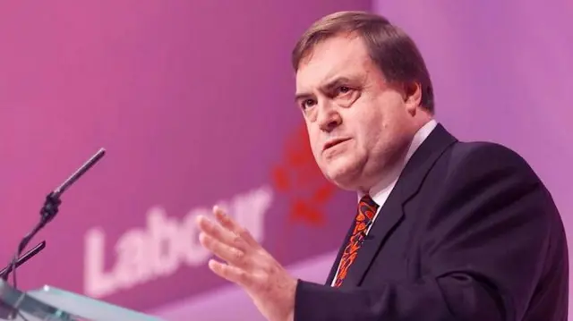 An old photo of Prescott gesturing with his hand as he speaks at a lectern in front of the Labour Party logo. He stands behind a lectern, his left hand raised. He's in a dark suit with a red tie and white shirt, a purple screen behind him with the words Labour and a red rose blurred on the wall